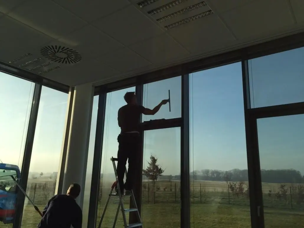 Glasreiniger auf einer Leiter vor einer raumhohen Fensterfront in einem Büro.