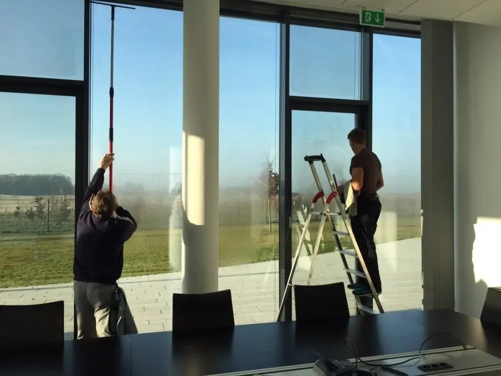 Zwei Glasreiniger arbeiten an einer raumhohen Fensterfront mit Leiter und Stange.