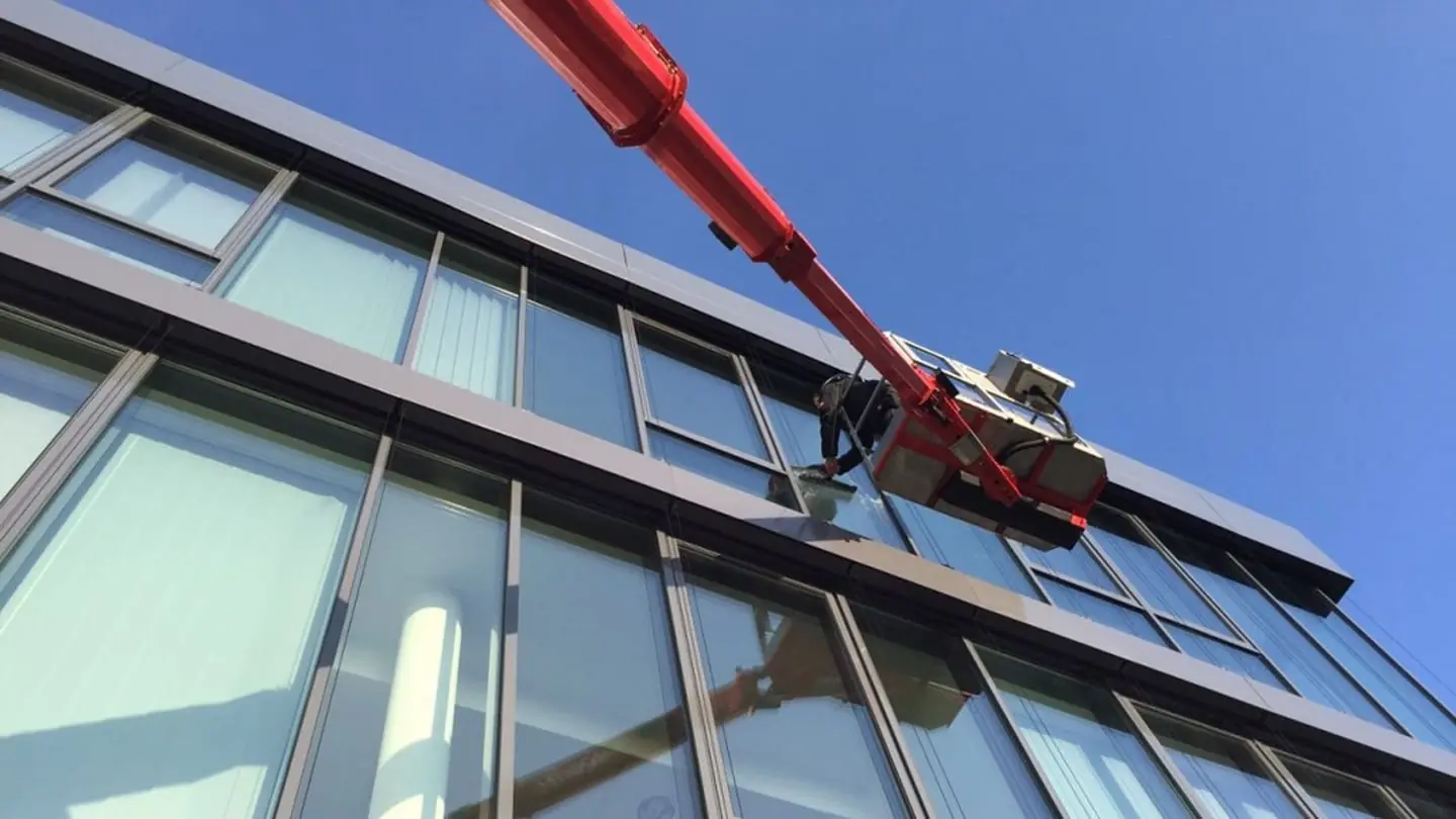 Reinigung einer Glasfassade mit einer roten Teleskopstange vor blauem Himmel.