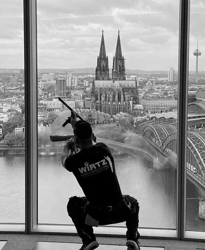 Ein WIRTZ-Glasreiniger kniet vor einer großen Fensterfront, dahinter der Kölner Dom.
