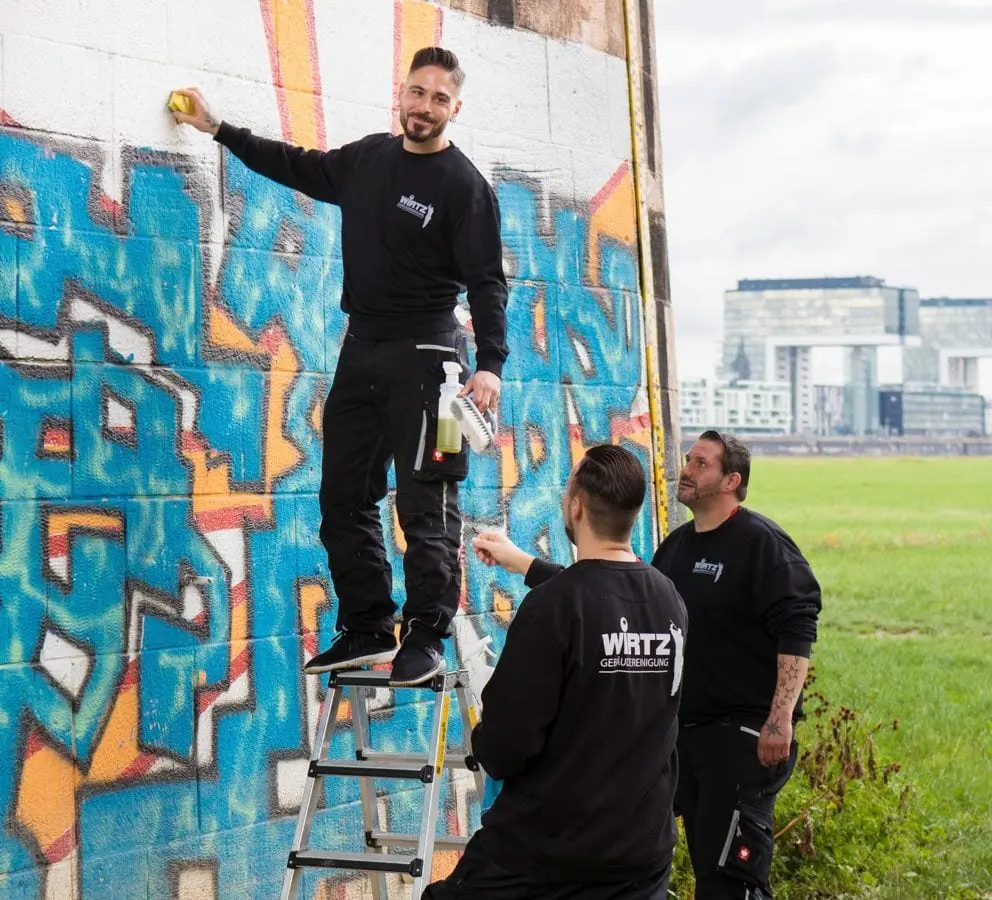 Zwei WIRTZ-Mitarbeiter entfernen ein großes Graffiti an einer Wand, im Hintergrund die Kranhäuser.