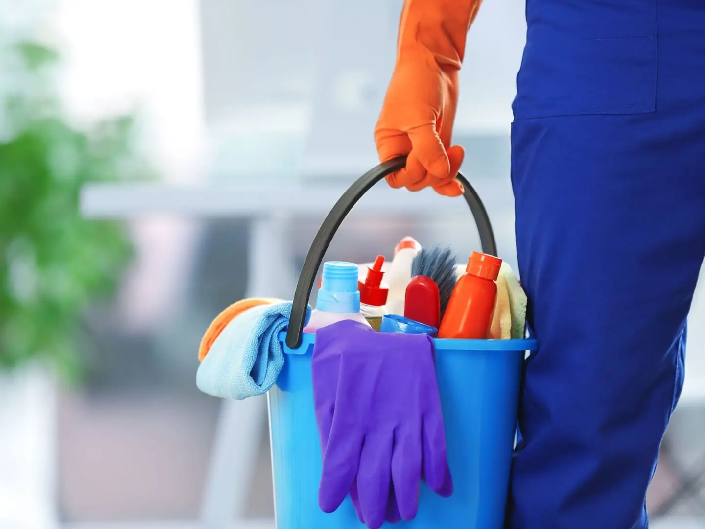 Eimer mit Reinigungsmitteln, Tüchern und Handschuhen in der Hand einer Reinigungskraft.