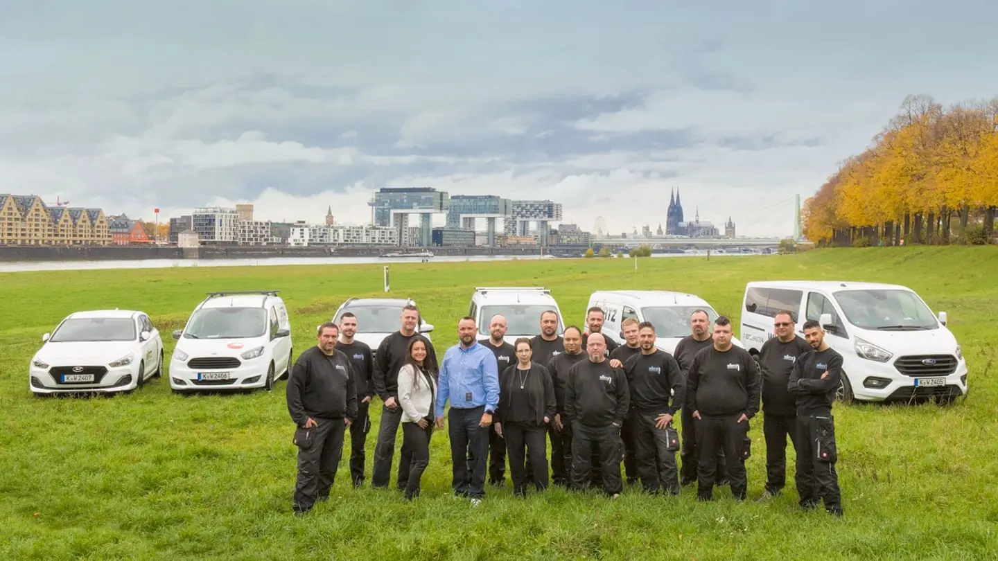 Das Team der WIRTZ Gebäudereinigung mit den Firmenwagen auf den Rheinwiesen, im Hintergrund die Kölner Kranhäuser und der Dom.